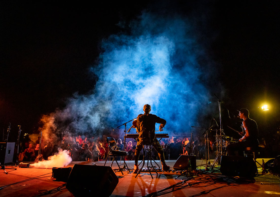 Foto von hinten auf eine Bühne, eine Band spielt. Im Hintergrund sind Menschen zu sehen. Eine Nebelmaschine stößt Nebel aus, der von Scheinwerfern bunt beleuchtet wird.